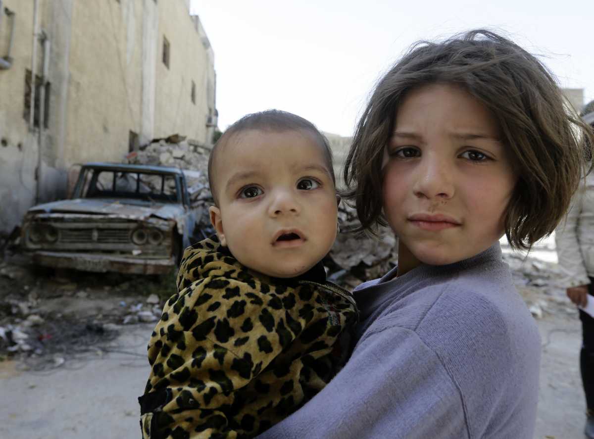 A picture taken during a Syrian army-organised tour on April 20, 2018 shows a girl holding a child while standing past destruction in a street in the Eastern Ghouta town of Douma on the outskirts of the capital Damascus. (Photo by LOUAI BESHARA / AFP)